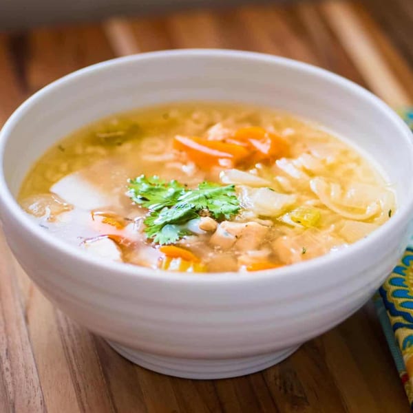 A white bowl of clear broth soup with chicken, vegetables, and cilantro on a wooden table.