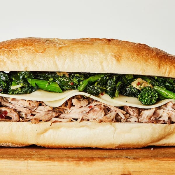 A large sandwich on a wooden board, filled with shredded meat, cheese, and green vegetables.