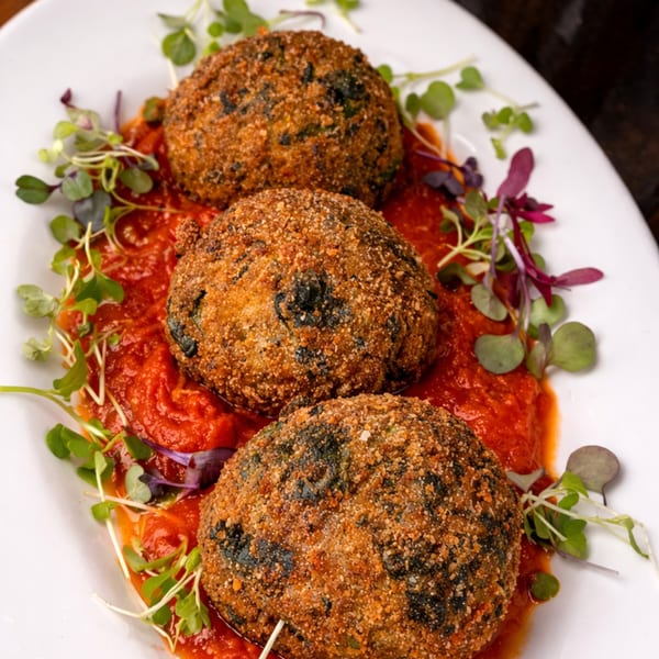 Three crispy, breaded arancini balls with dark fillings, served on a bed of red tomato sauce and microgreens.